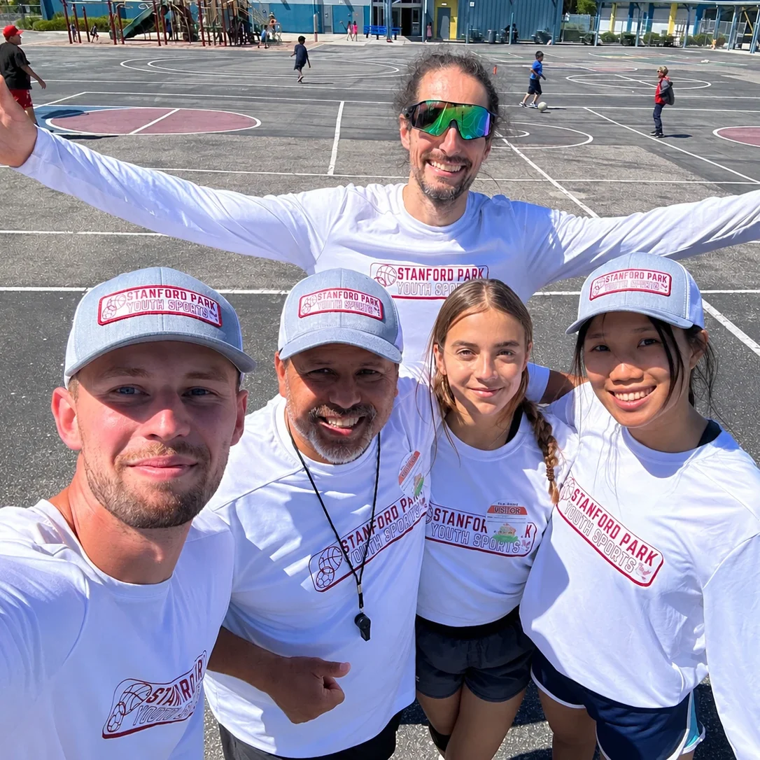Five smiling Stanford Park Youth Sports coaches in matching caps and shirts on a school blacktop